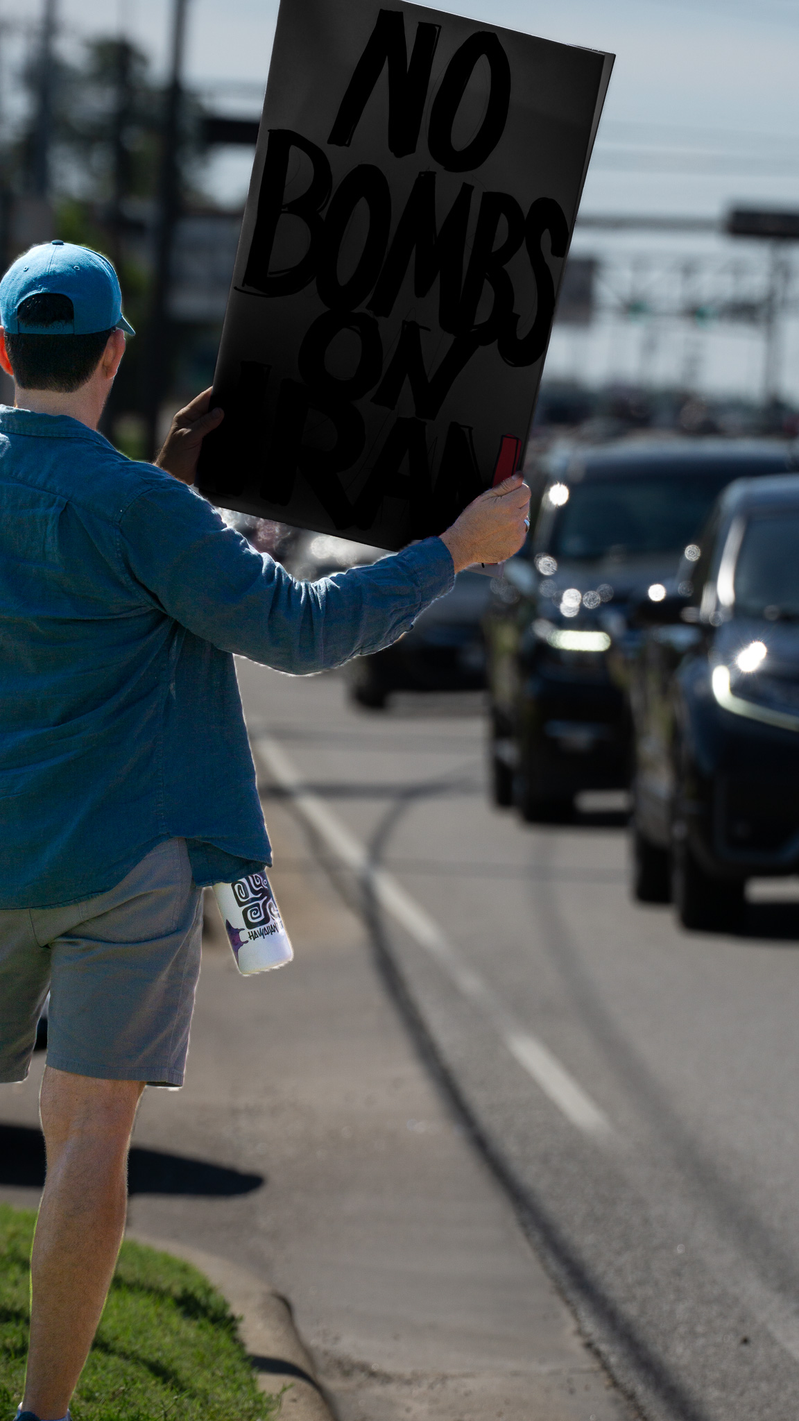 Artistic edit: person holding No Bombs on Iran sign in traffic