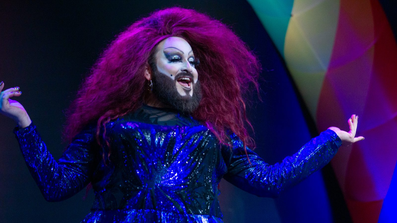 Drag performer with a magenta wig and blue sequined gown under stage light