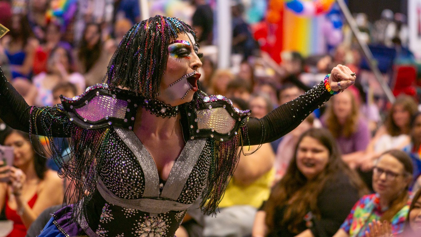 Drag performer in a rainbow-beaded outfit with arms spread mid-performance