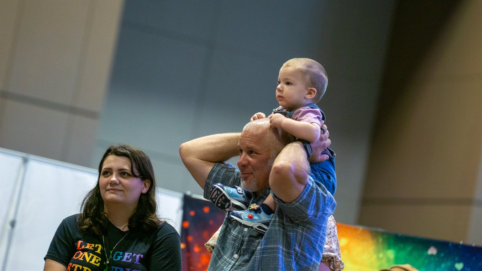 Toddler watching the show from a parent's shoulders in front of a rainbow banner at Pride 2026